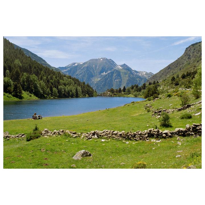 A couple sits at a picnic table by a lake surrounded by mountains and forest.