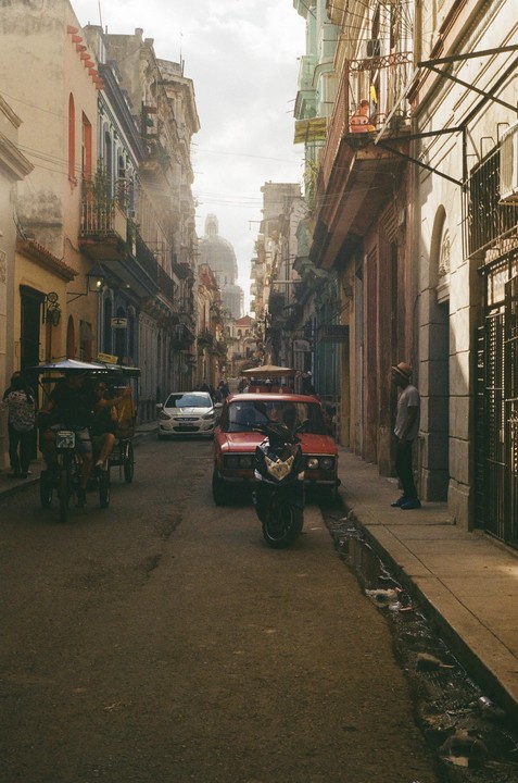 A narrow street in Cuba is filled with pedestrians, a red car, a motorcycle, and a bicycle rickshaw, with a large dome building visible in the distance.