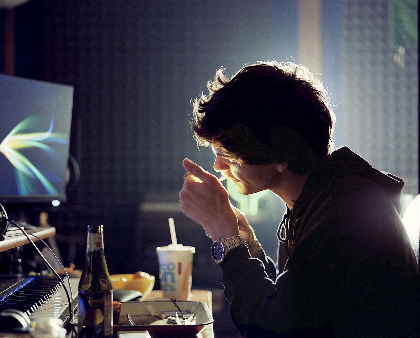A man lights a cigarette at a desk with a computer monitor, keyboard, and drinks.