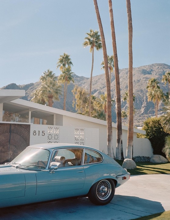 A light blue vintage car is parked in front of a white mid-century modern house with palm trees and mountains in the background.