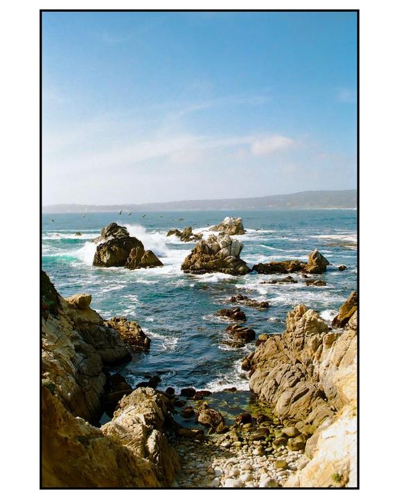 Waves crash against rocky outcroppings along a coastline under a clear blue sky.