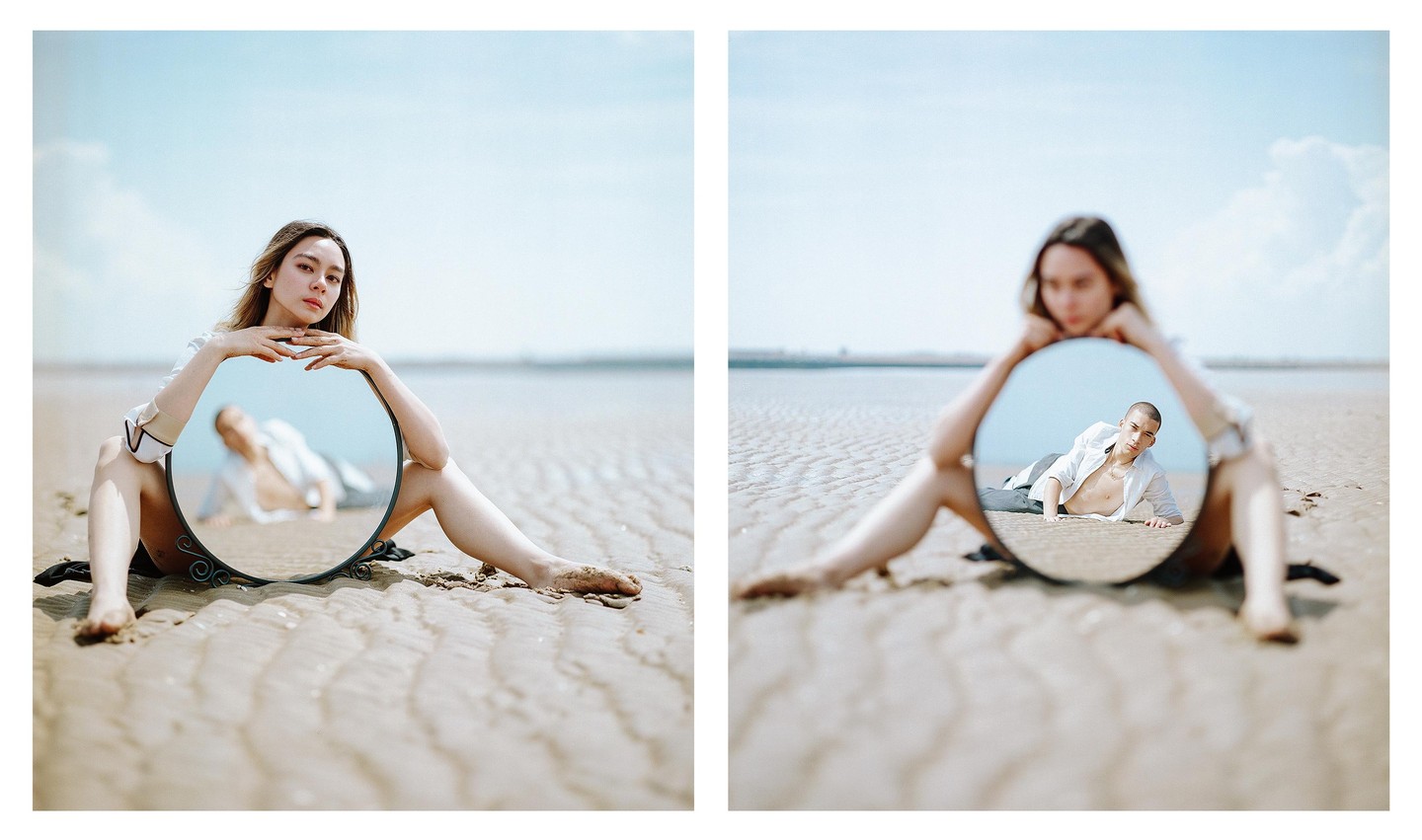 A woman sits on a sandy beach holding a round mirror that reflects a man lying on the sand.