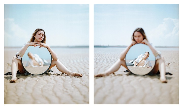A woman sits on a sandy beach holding a round mirror that reflects a man lying on the sand.