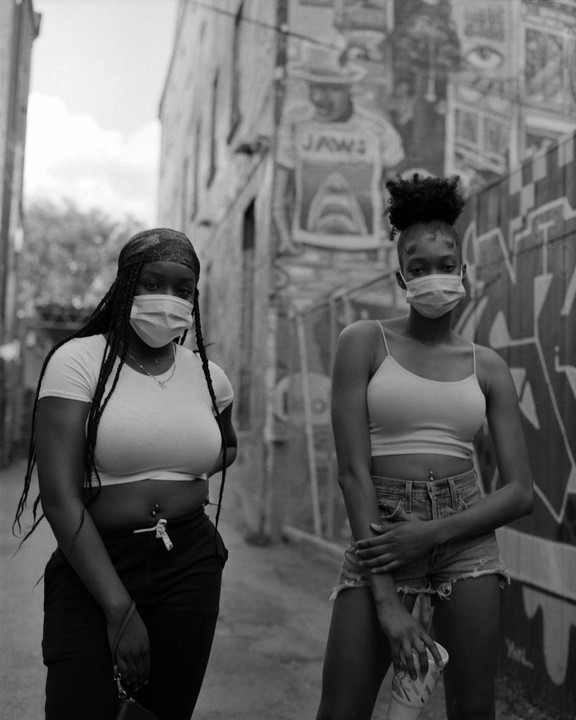 Two women wearing face masks stand in an alley with graffiti and a mural on the wall.