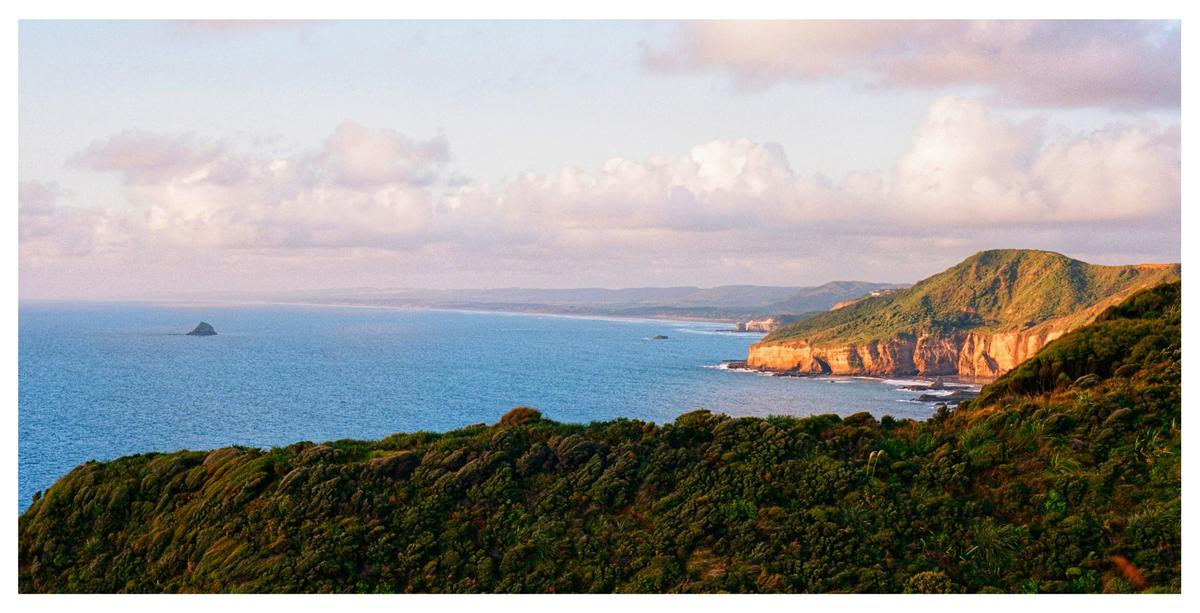 A panoramic landscape shows a rugged coastline with green hills, cliffs, and a small island in the ocean under a cloudy sky.