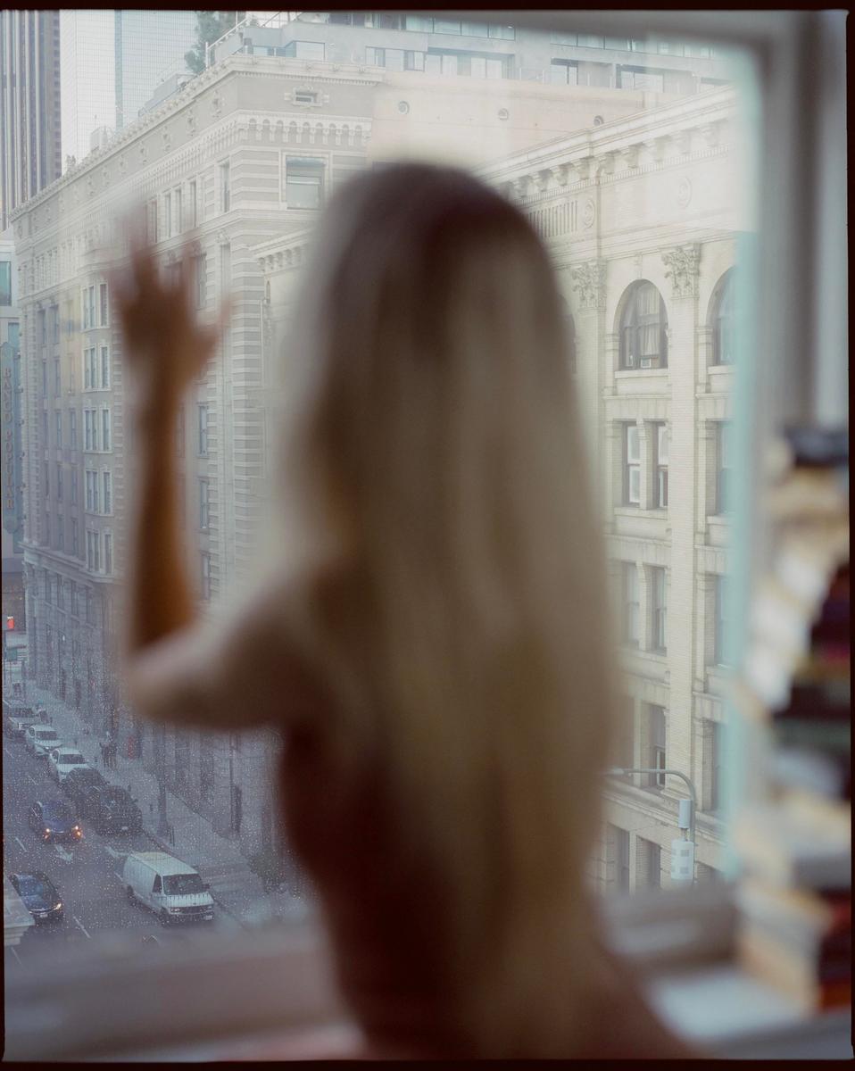 A woman with long blonde hair looks out a window at a city street and buildings.