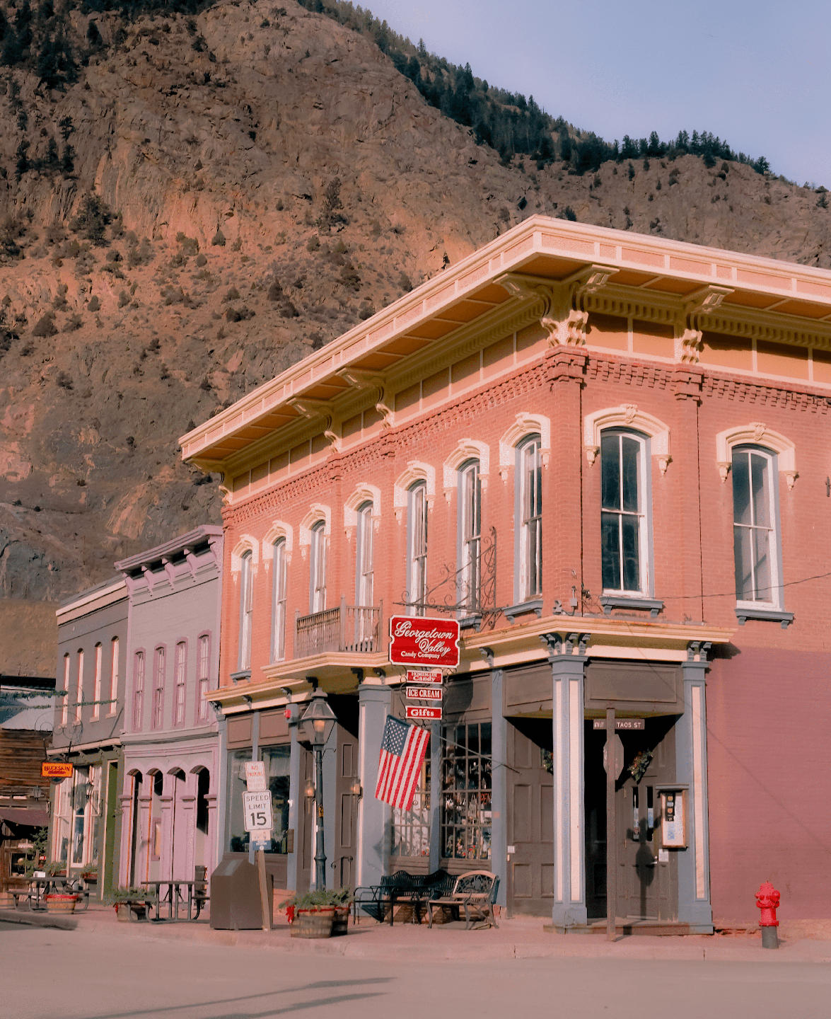 A brick building with an American flag hanging from its facade stands on a street in front of a mountain.