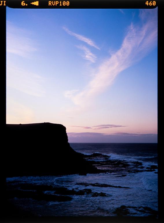 A dark cliff face is silhouetted against a colorful sky over a rocky ocean.