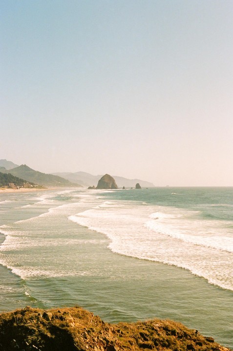 Waves roll onto a beach with a large rock formation in the distance and mountains along the coast.