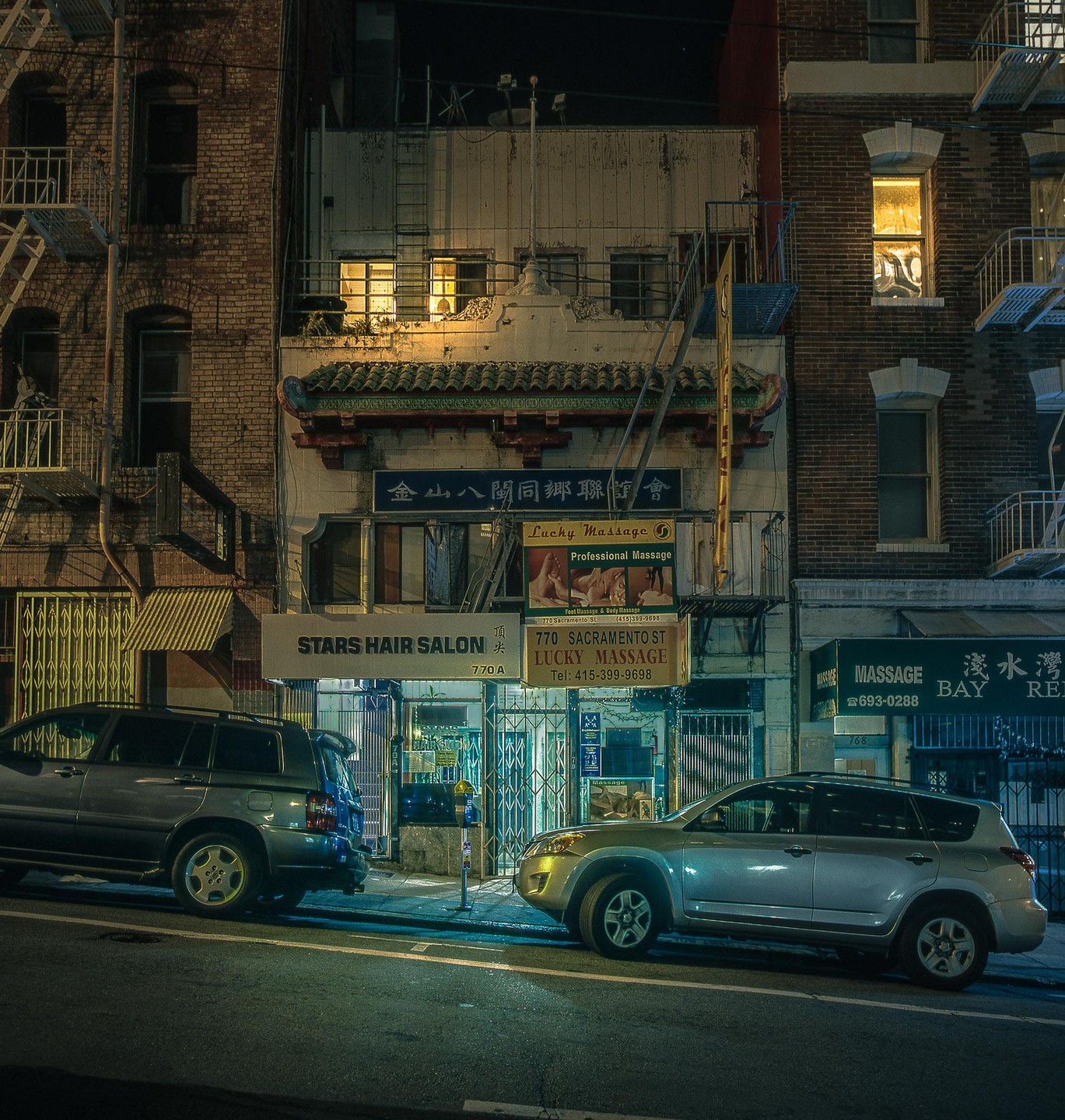 Two cars are parked on a street in front of buildings with businesses at night.