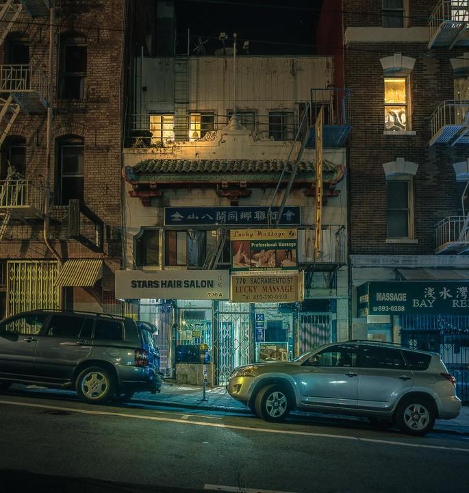 Two cars are parked on a street in front of buildings with businesses at night.