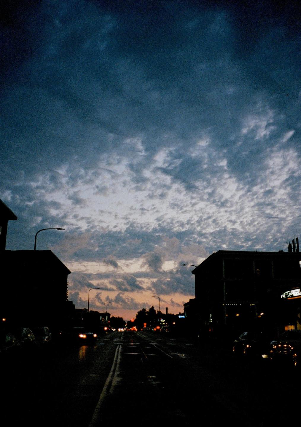 A street scene at dusk shows buildings silhouetted against a cloudy sky with car lights visible in the distance.