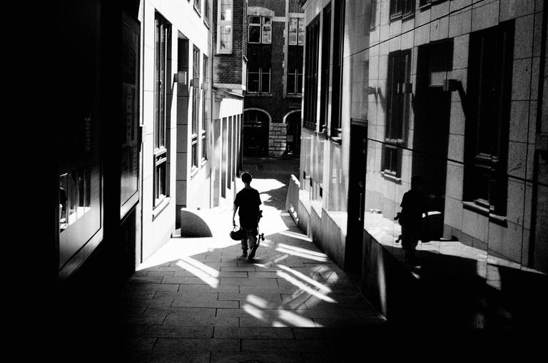 A boy walks down a narrow alleyway between tall buildings, carrying a skateboard and a helmet.