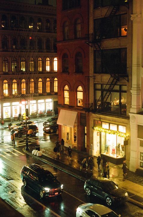 A street in SoHo is wet from rain, with cars and people moving past brightly lit buildings at night.