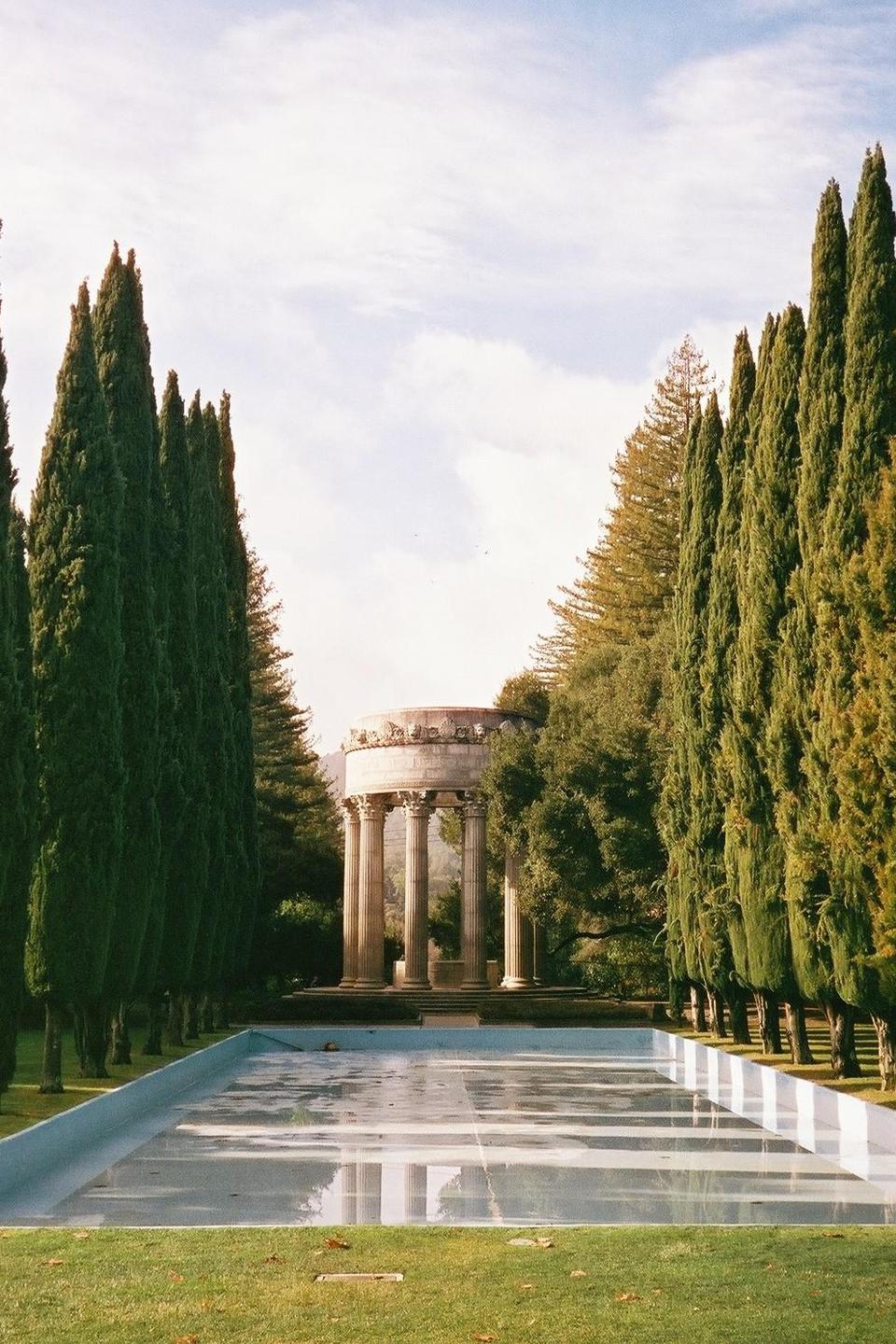A classical rotunda with columns stands at the end of a reflecting pool, flanked by tall cypress trees.