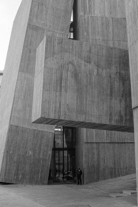 A man stands in front of a brutalist concrete building with geometric shapes and glass windows.