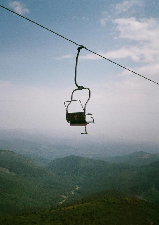 A ski lift chair hangs over a vast mountain landscape under a cloudy sky.