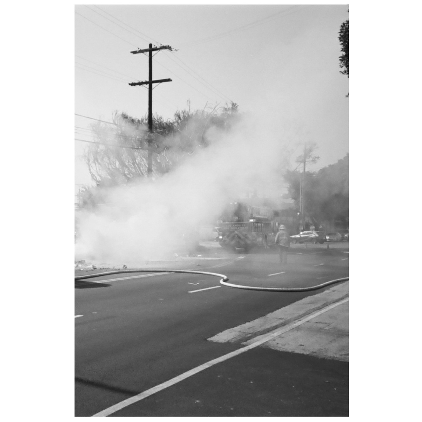 A firefighter stands in the street with a fire truck and smoke in the background.