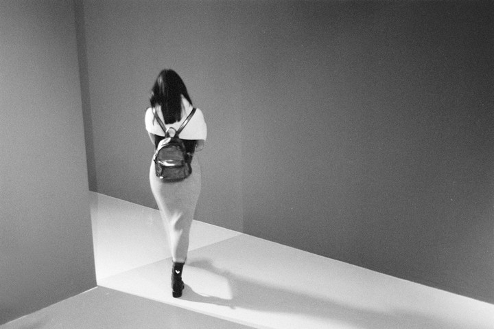 A woman with long hair and a backpack walks away from the camera down a hallway with strong shadows.