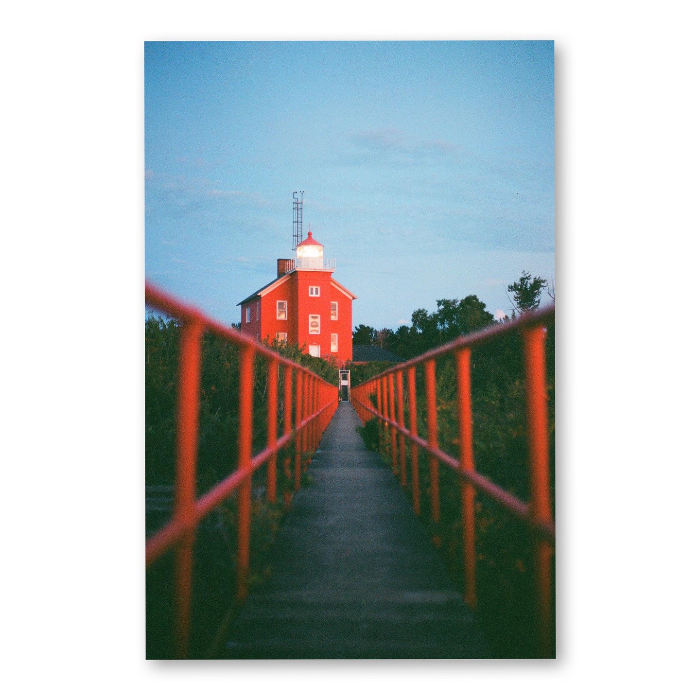 A red lighthouse with its light on stands at the end of a long walkway with red railings, surrounded by green foliage under a clear sky.