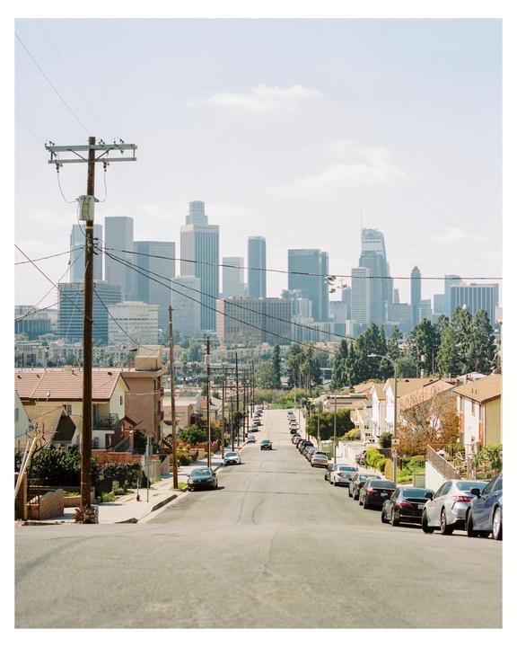 A long street with parked cars on both sides leads down a hill toward a distant city skyline.