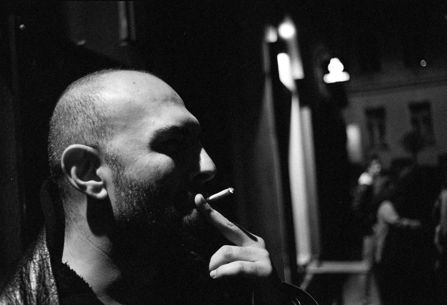 A man with a beard and shaved head smokes a cigarette in profile, with a dark background and blurred figures.