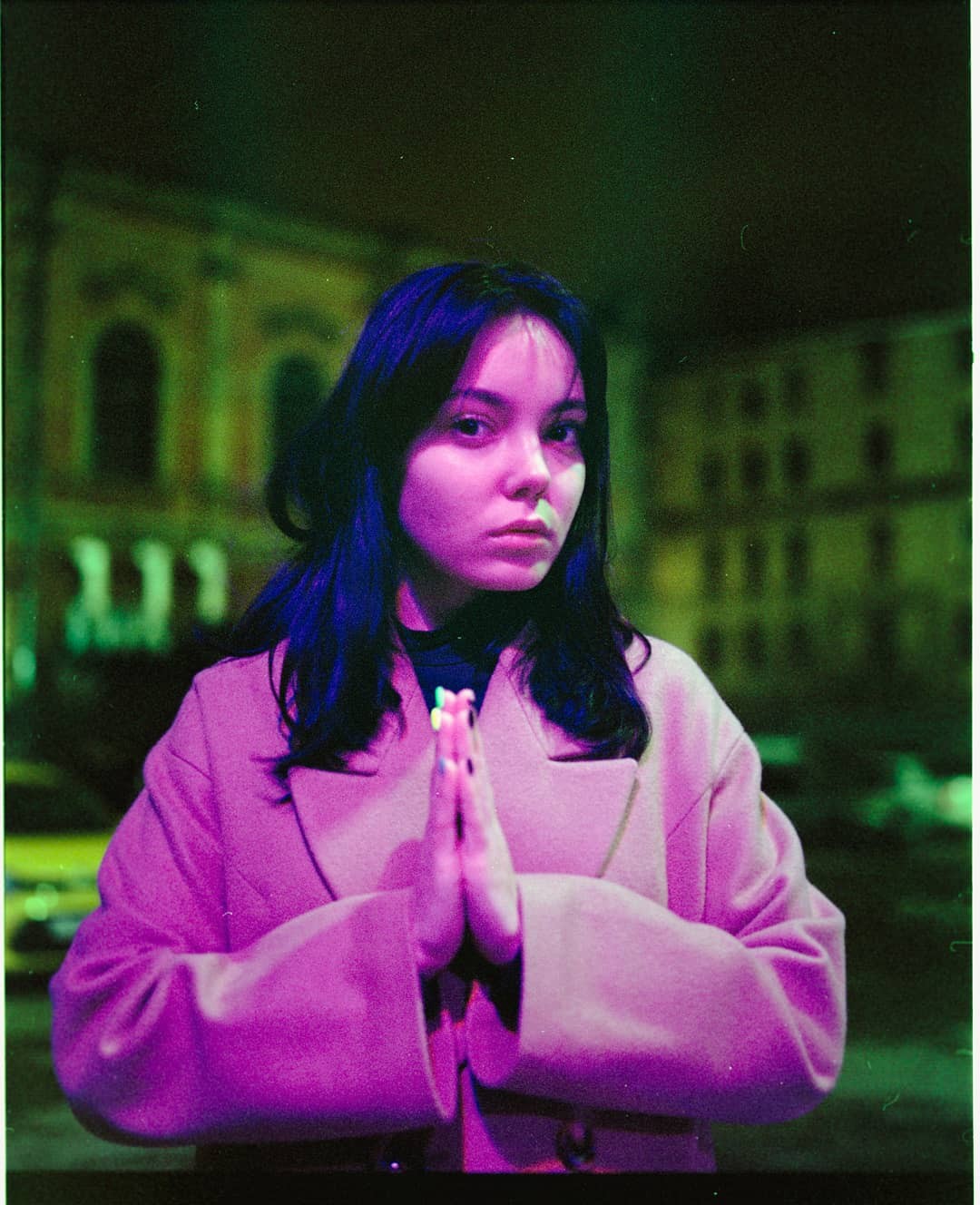A woman in a pink coat stands with her hands pressed together in front of a building at night.