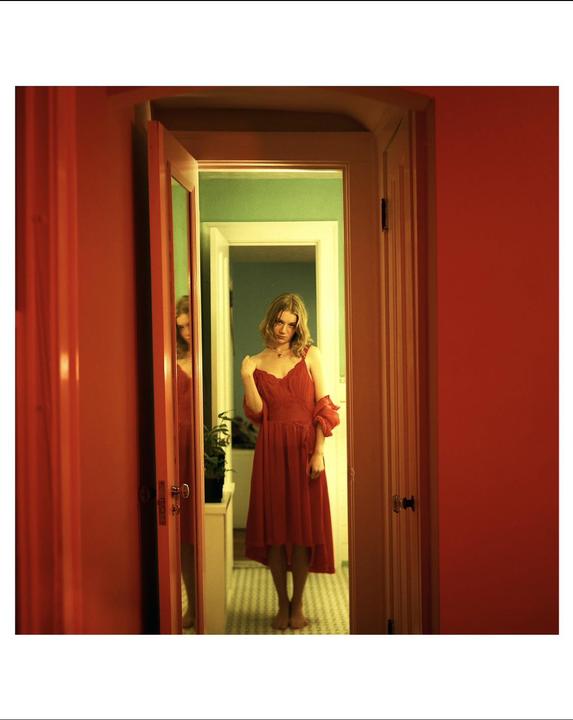 A woman in a red dress stands barefoot in a doorway, with a reflection visible in a mirror to her left.