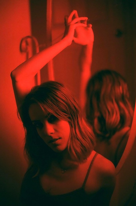 A woman with short hair looks at the viewer, with her reflection visible in a mirror behind her, all bathed in red light.
