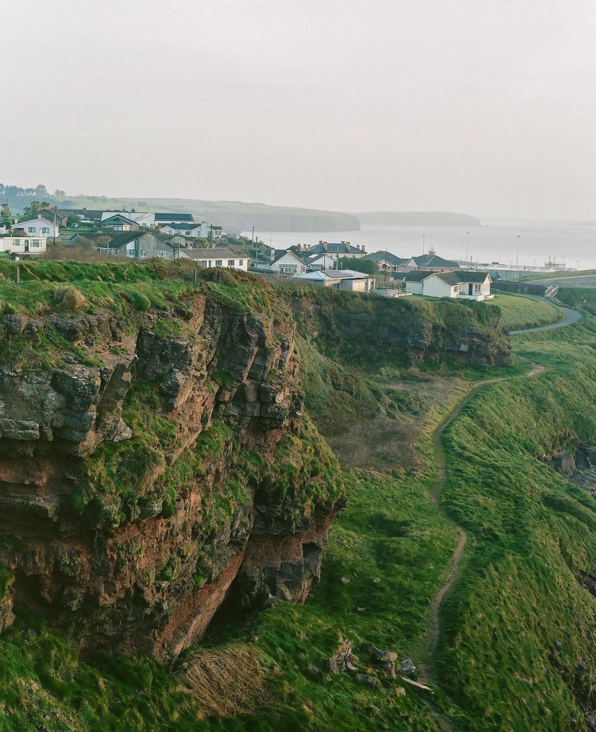 A small village sits atop a cliff overlooking the sea, with a winding path leading down the grassy hillside.