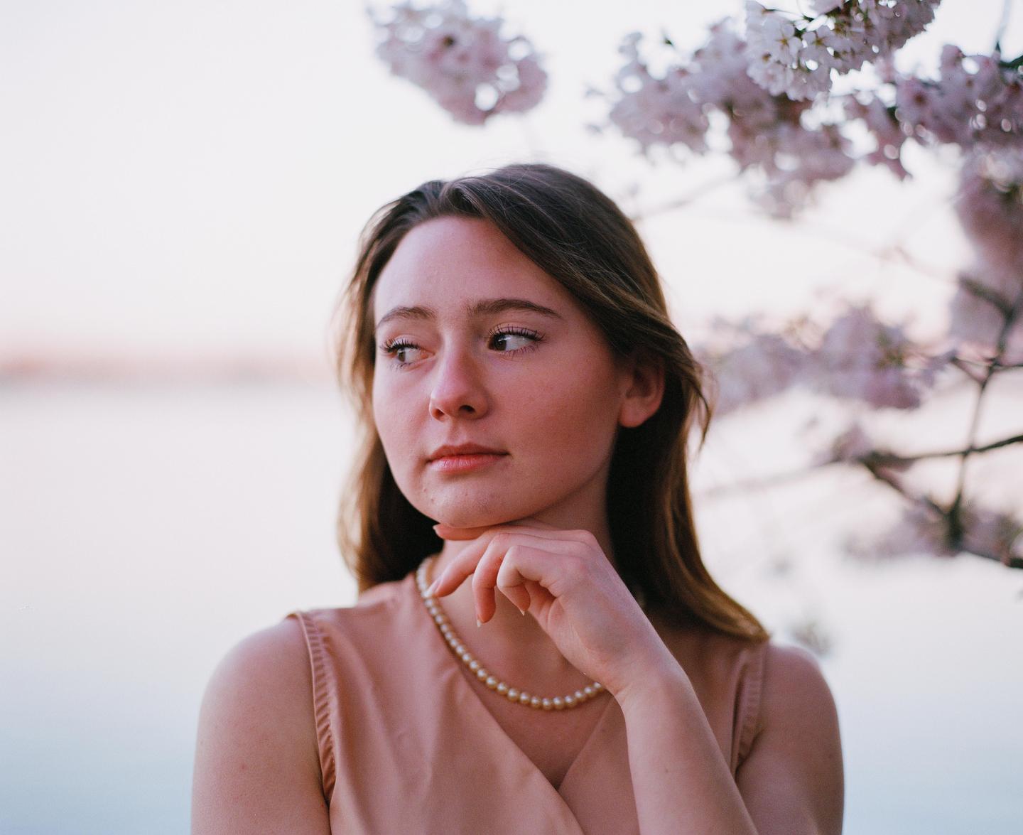 A young woman wearing a pearl necklace looks away from the camera with cherry blossoms in the background.
