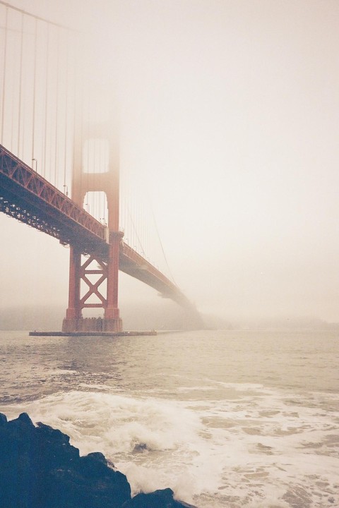 The Golden Gate Bridge is partially obscured by fog, with waves crashing on rocks in the foreground.