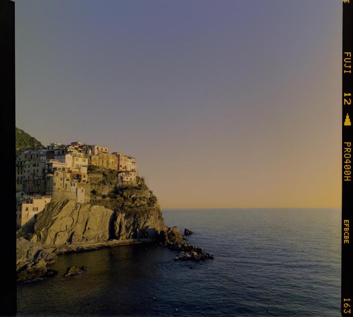 A village with colorful buildings is perched on a rocky cliff overlooking the sea at sunset.
