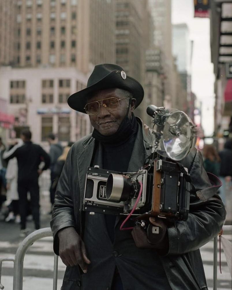 A man wearing a hat and sunglasses holds a large format camera with a flash attached to it on a city street.