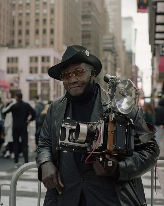 A man wearing a hat and sunglasses holds a large format camera with a flash attached to it on a city street.