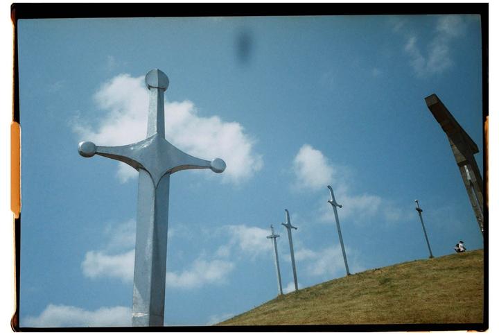 Several large sword sculptures are planted in a grassy hill under a blue sky with white clouds.