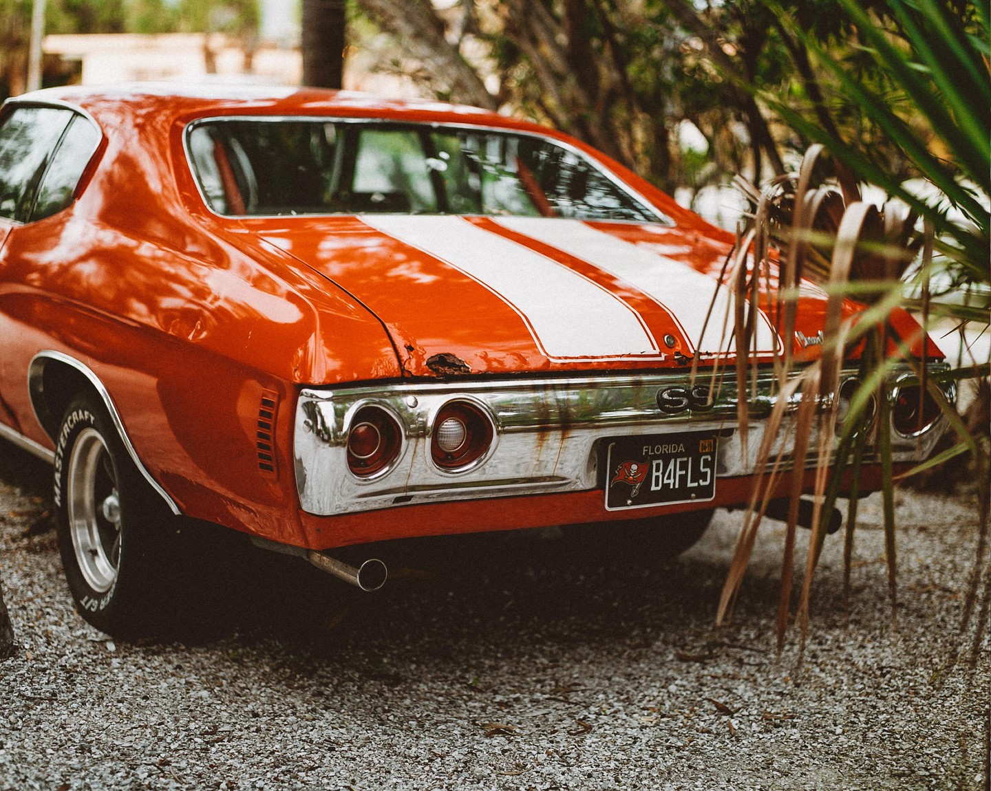 The rear of a vintage orange car with white racing stripes is parked on a gravel surface, partially obscured by green foliage.