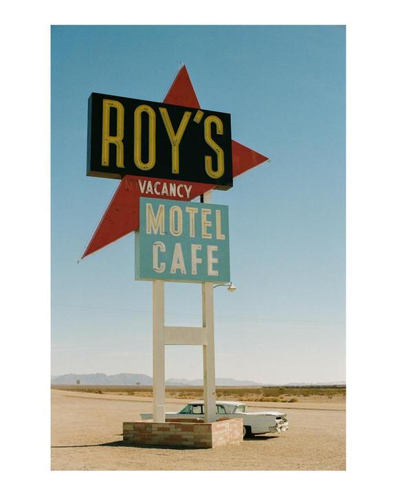 A large sign for Roy's Motel and Cafe stands in a desert landscape with a vintage car parked nearby.