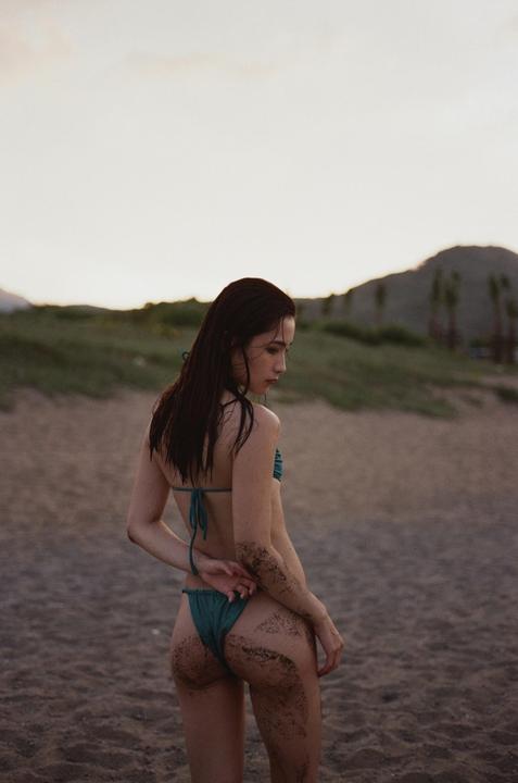 A woman in a bikini stands on a sandy beach with mountains in the background.