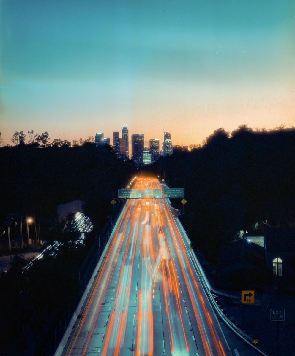 A highway with light trails from cars leads to a city skyline at dusk.