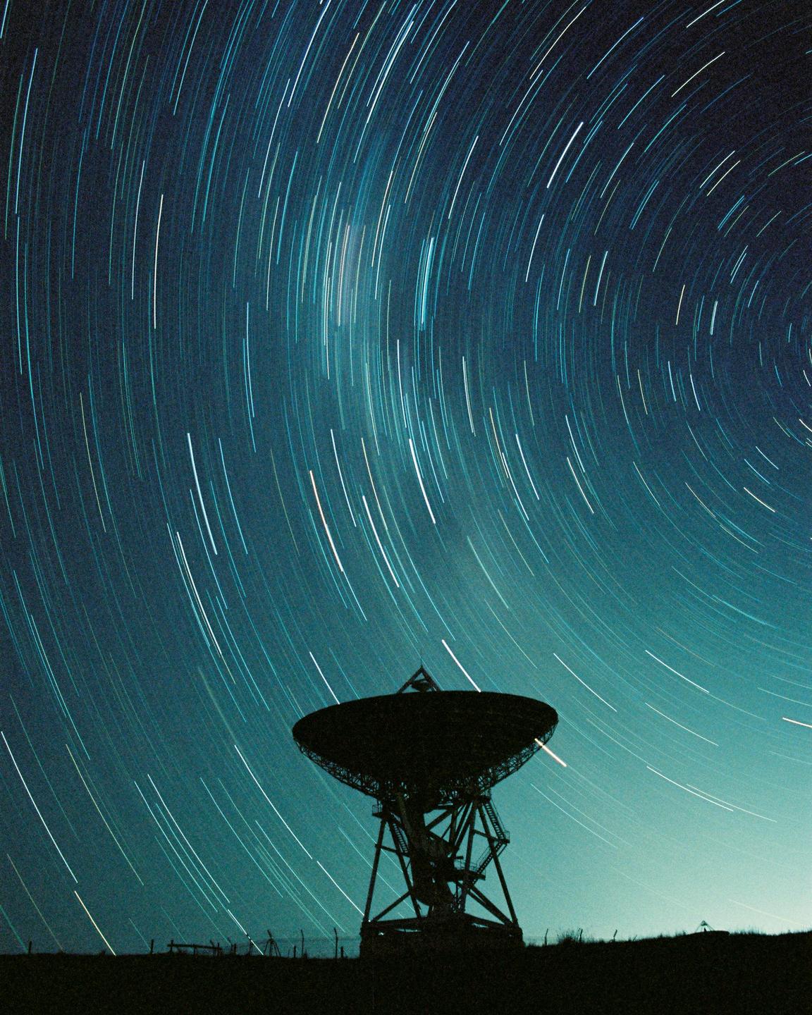 A radio telescope is silhouetted against a night sky filled with star trails.