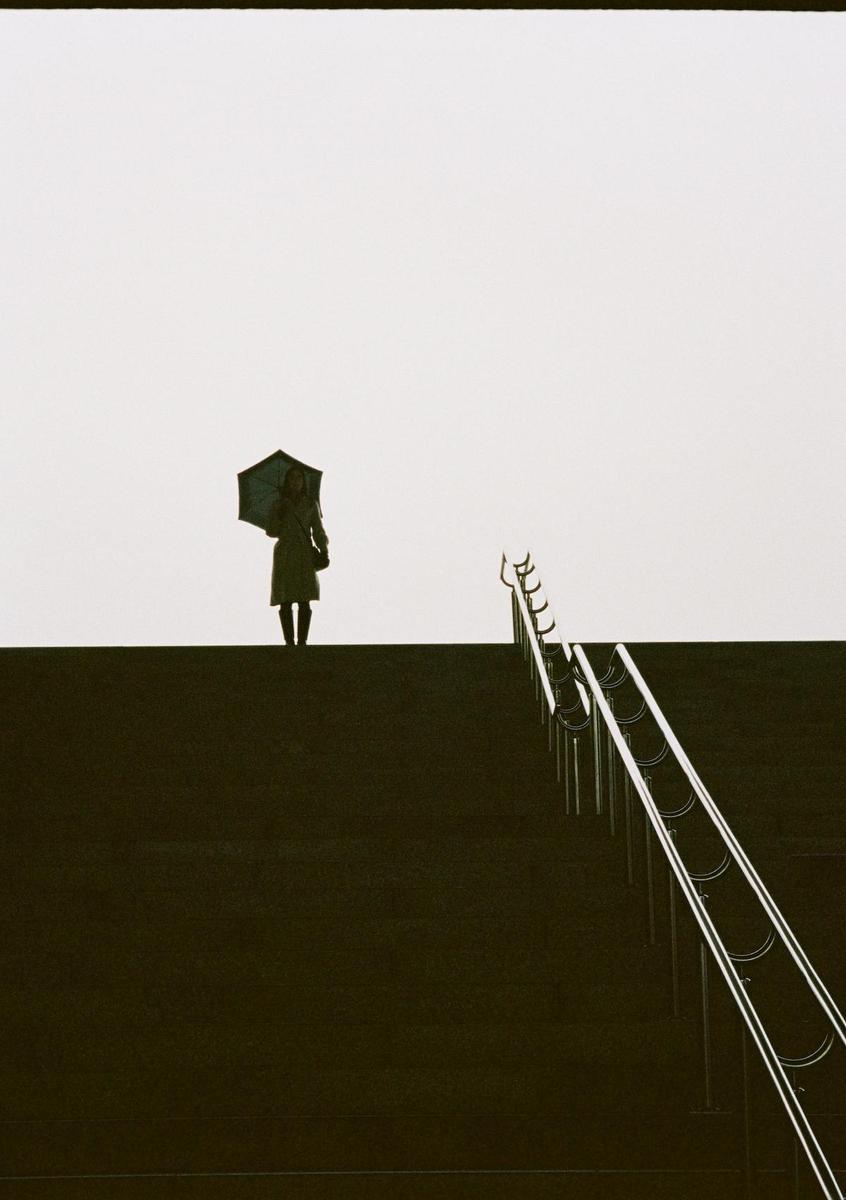 A woman holding an umbrella stands on a dark elevated surface next to a railing.