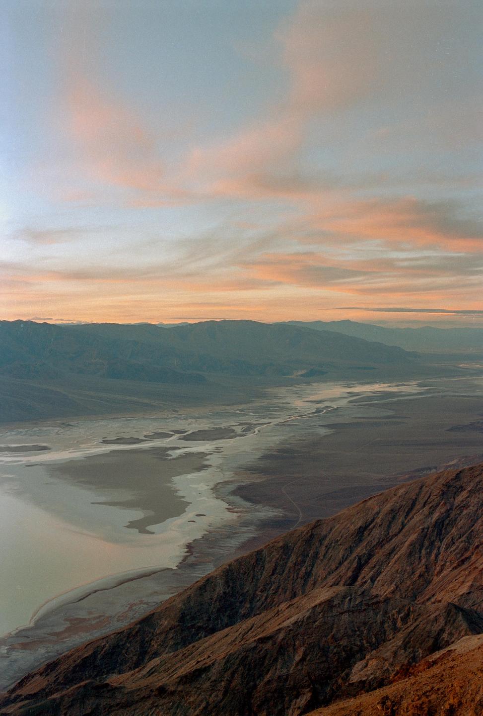 A vast desert landscape with mountains and a salt flat is visible under a sky with pink clouds at sunset.