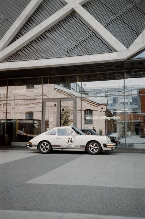 A white Porsche with the number 74 on its side is parked in front of a modern building with large glass windows.