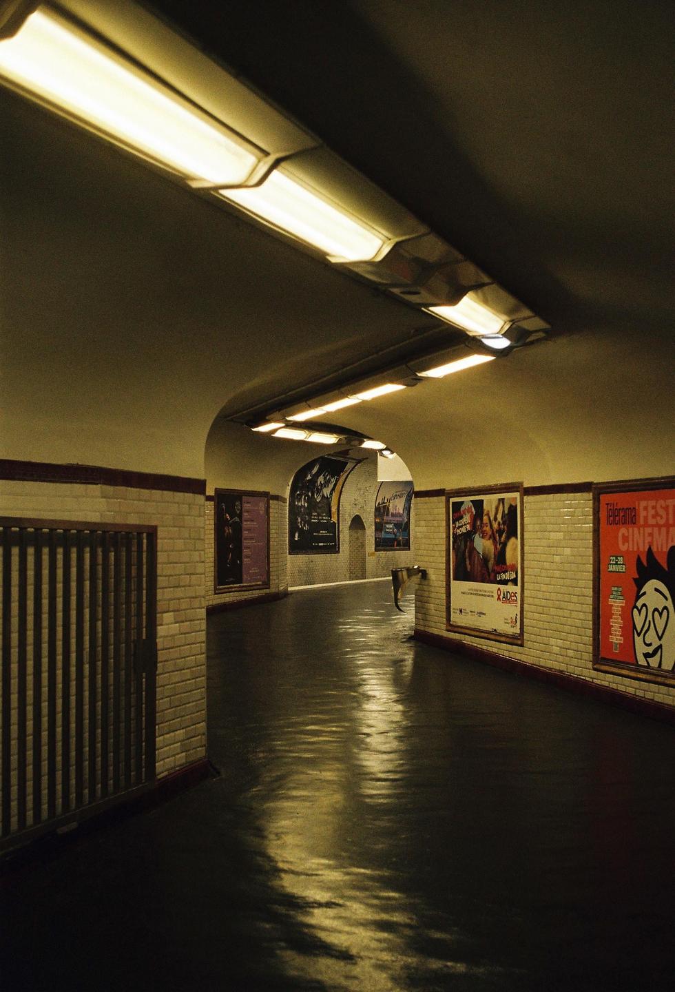 A long, curved underground tunnel is illuminated by fluorescent lights, with posters on the tiled walls and reflections on the wet floor.