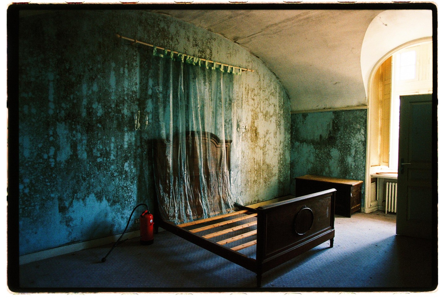 An empty wooden bed frame sits in a dilapidated room with peeling blue wallpaper and a fire extinguisher on the floor.