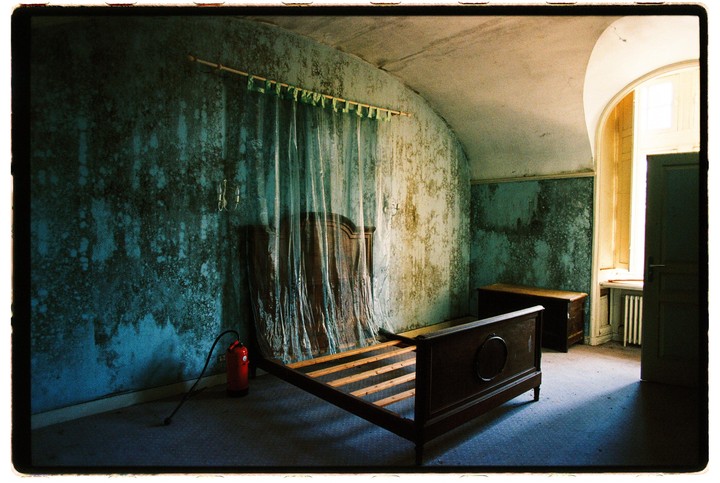An empty wooden bed frame sits in a dilapidated room with peeling blue wallpaper and a fire extinguisher on the floor.