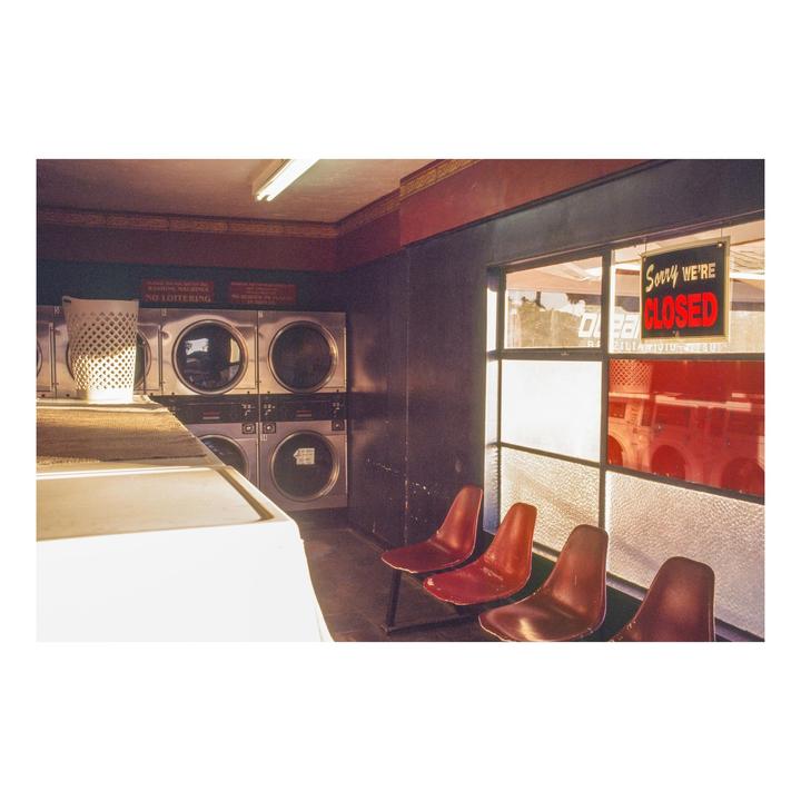 A laundromat interior features washing machines, red chairs, and a "Sorry We're Closed" sign in the window.