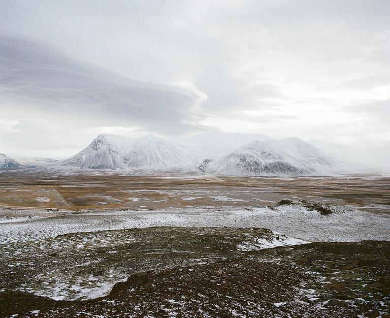 A vast, snow-dusted landscape stretches towards snow-capped mountains under a cloudy sky.
