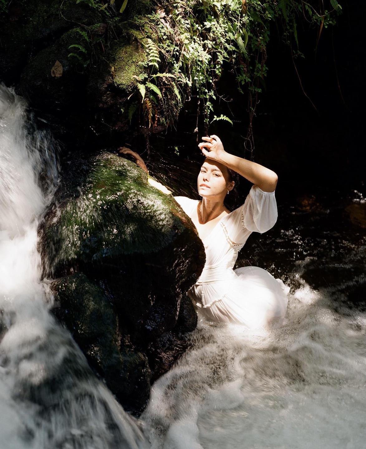 A woman in a white dress stands in a waterfall with sunlight on her face.
